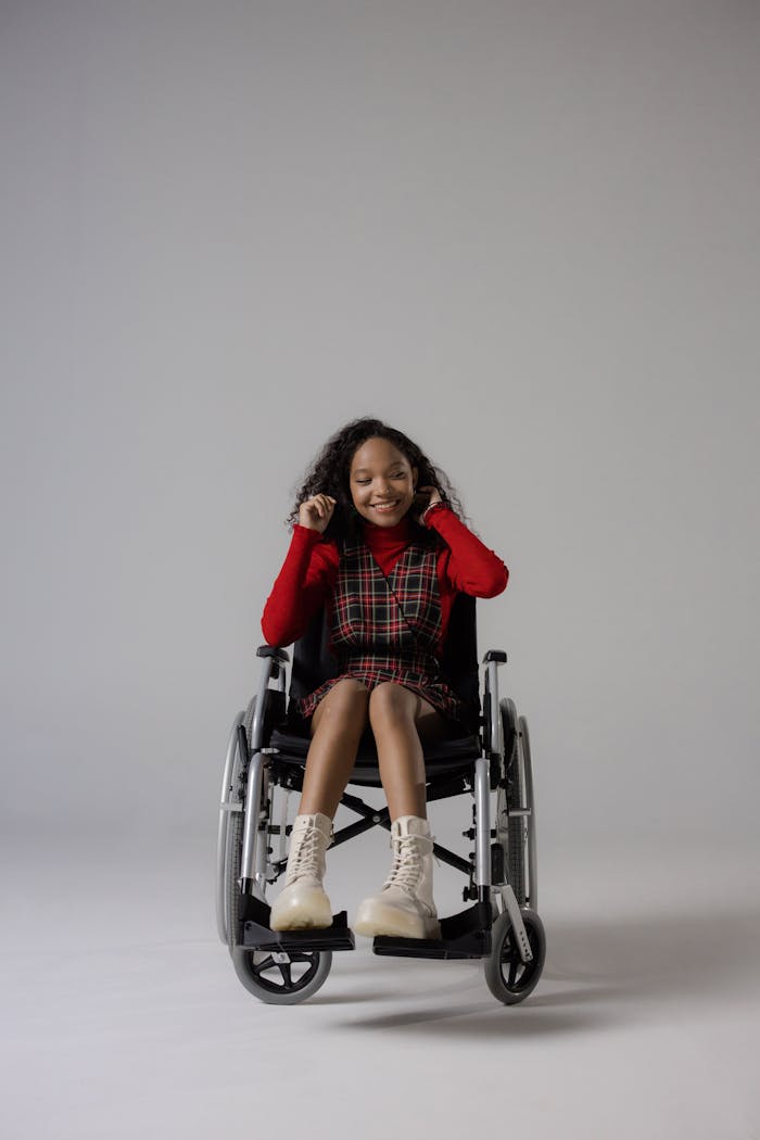 A cheerful young woman with curly hair in a stylish outfit sitting in a wheelchair against a gray studio background.